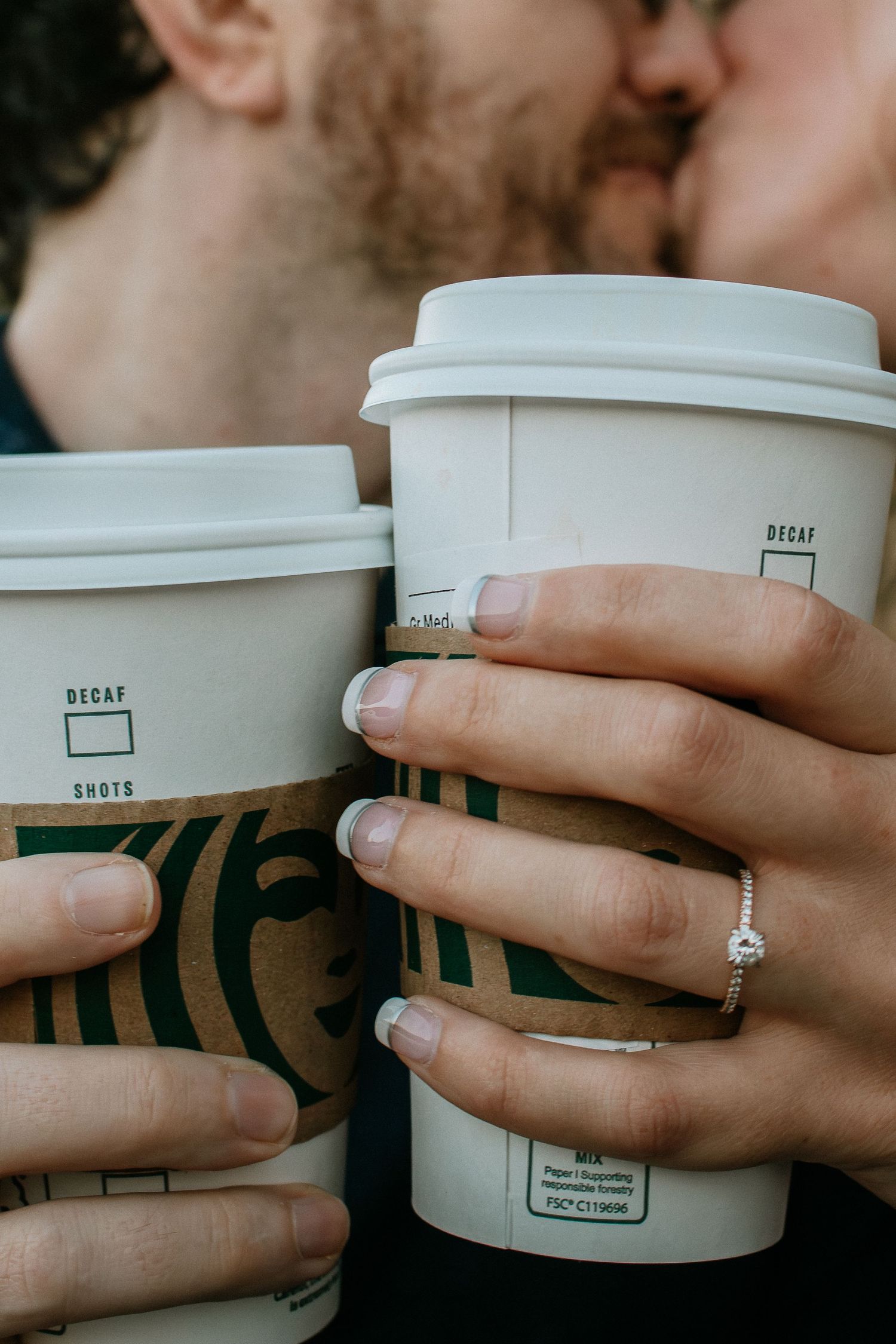 Couple kissing behind two Starbucks cups, one hand showing a diamond engagement ring at Winton Woods in Cincinnati, Ohio