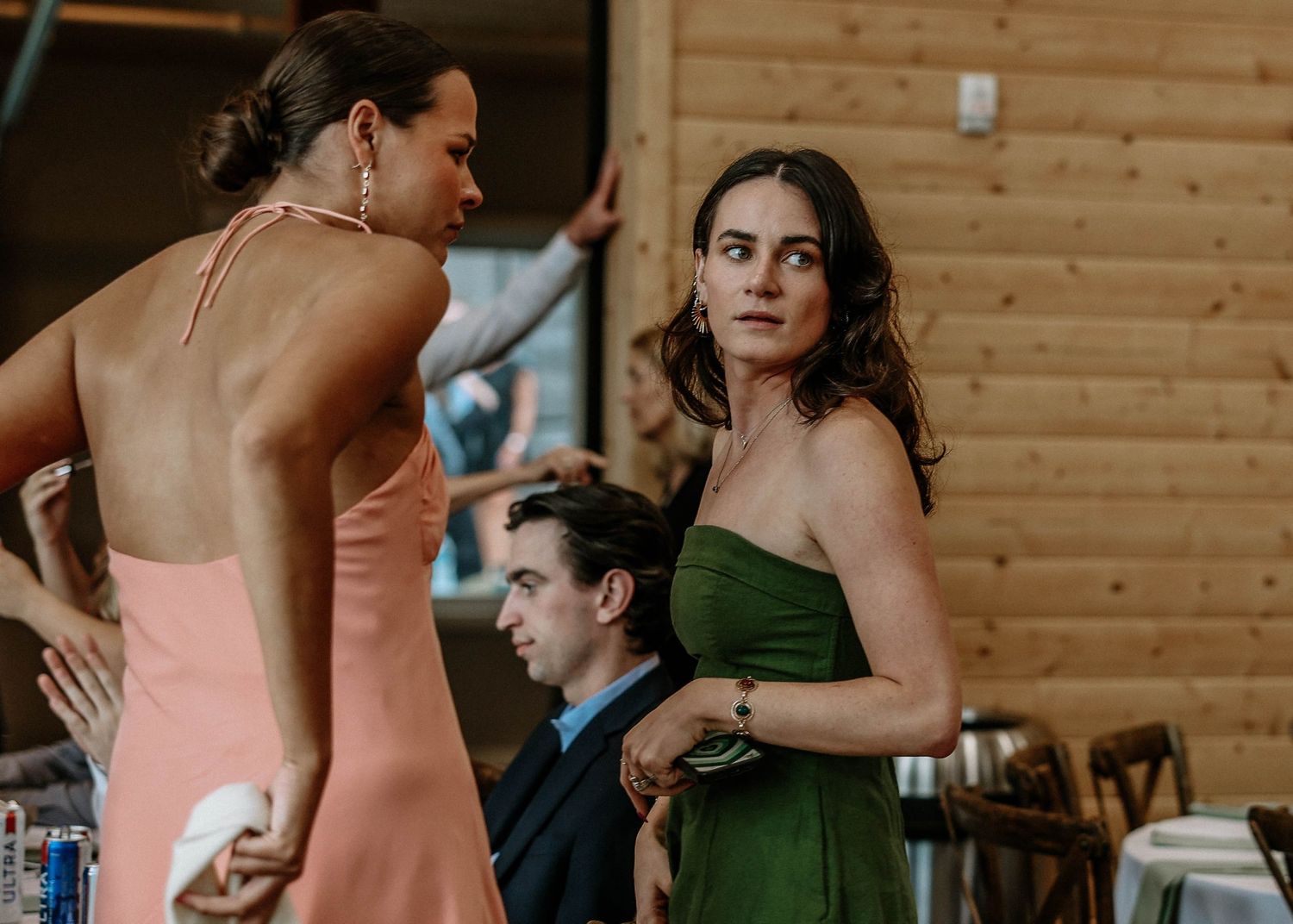 Two women in elegant dresses, one pink and one green, converse at a formal indoor event with wooden walls.