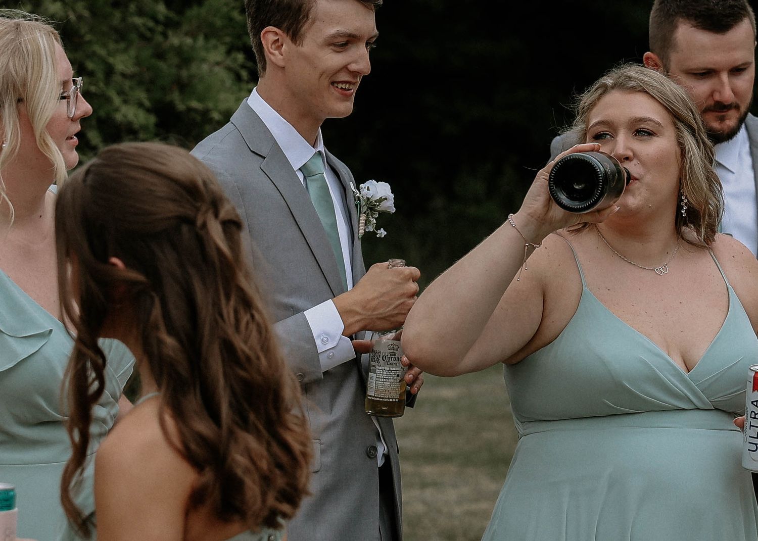 Bridesmaid in sage green gown drinks from a bottle while groomsman in gray suit smiles at outdoor wedding.