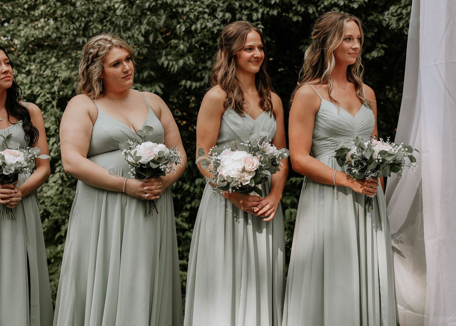 Bridesmaids in sage green gowns holding white floral bouquets stand together during an outdoor wedding ceremony.