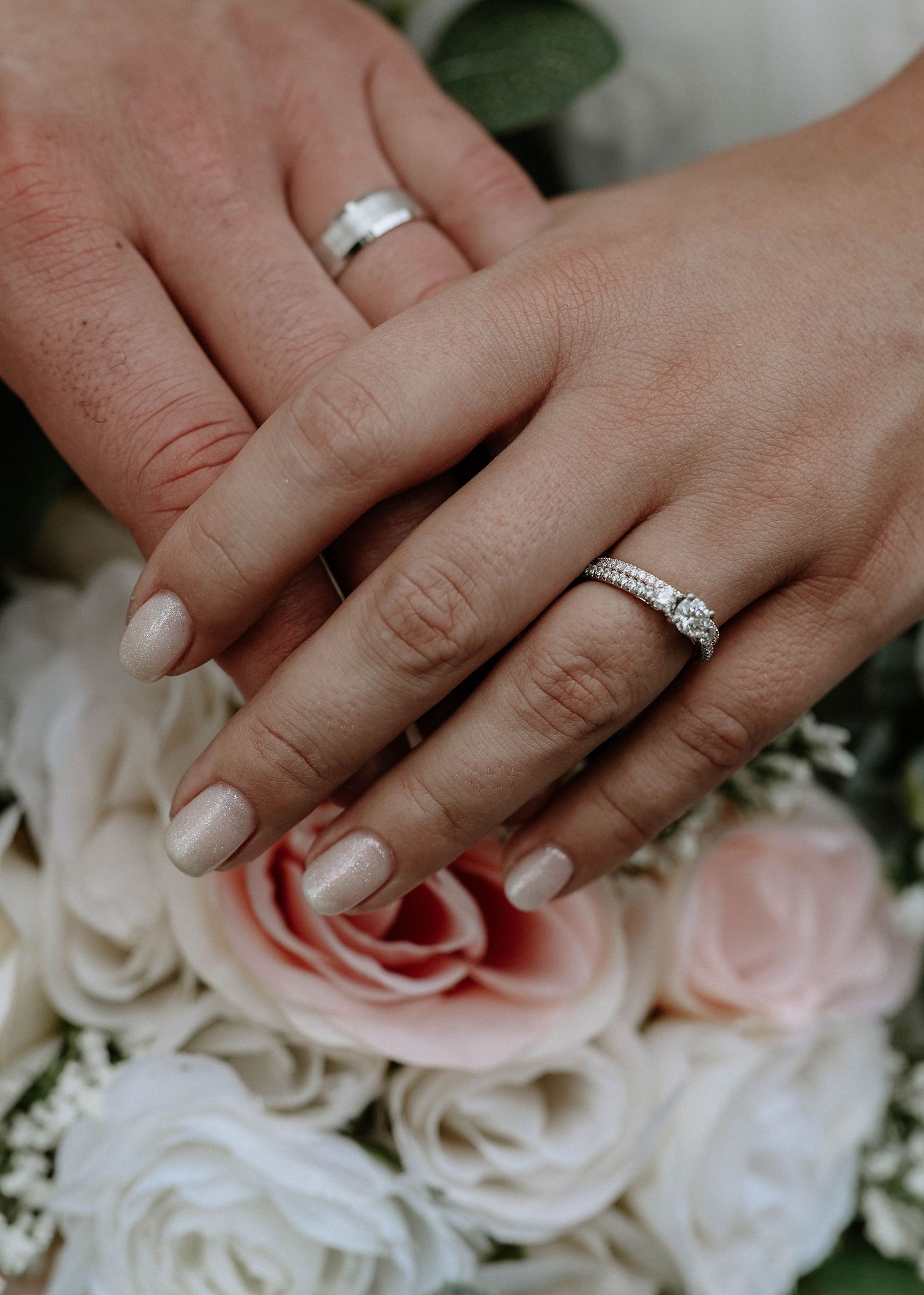 Two hands with wedding rings resting on pink and white roses, symbolizing marriage and love.