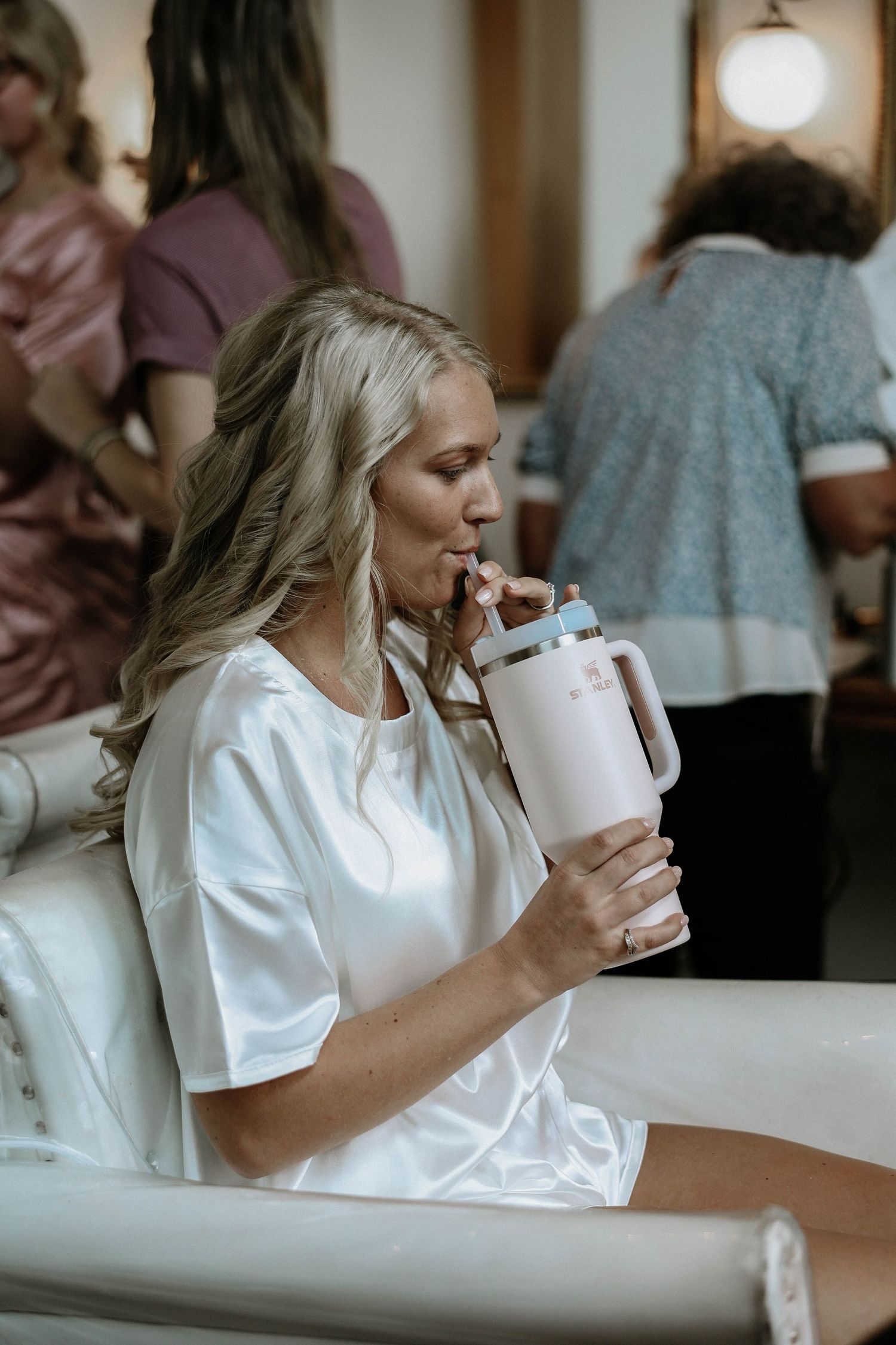A blonde bride in a white satin robe sipping from a pink tumbler while getting ready for her wedding.