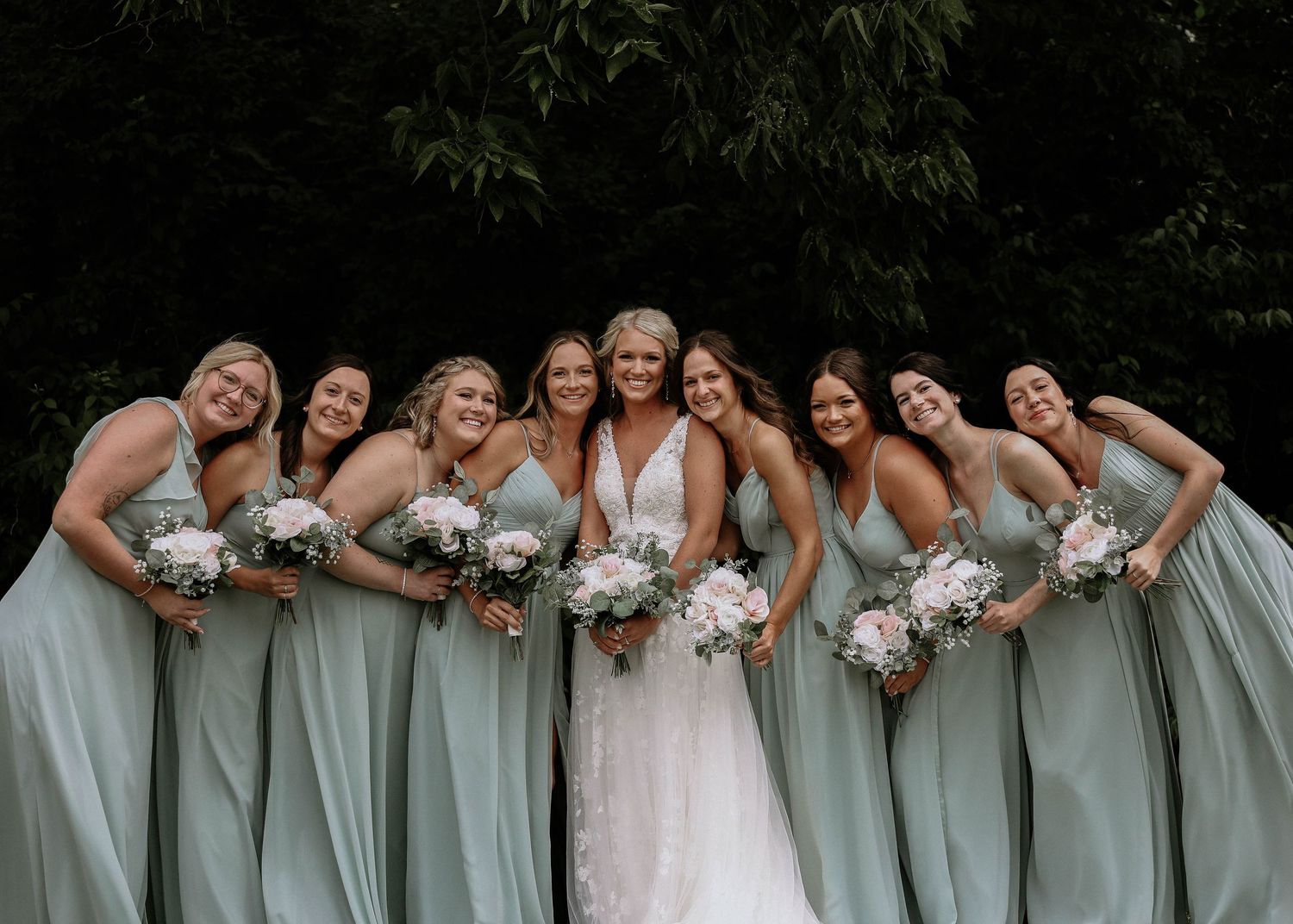 Bride surrounded by smiling bridesmaids in sage green gowns holding pink and white bouquets.