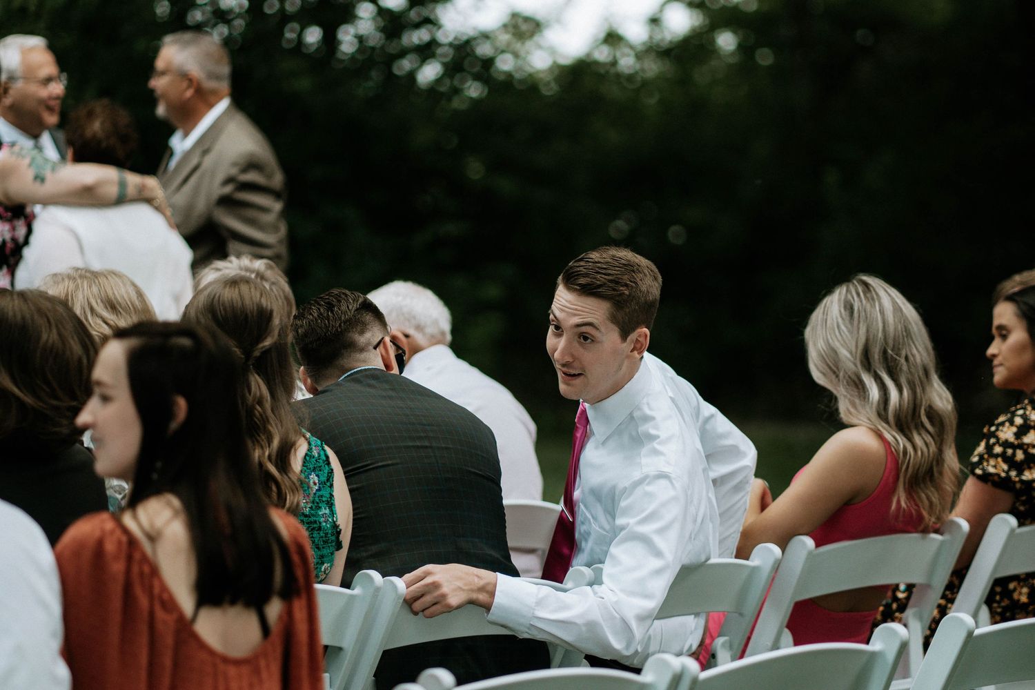 Young man in white shirt greeting guests at outdoor wedding ceremony seating area.