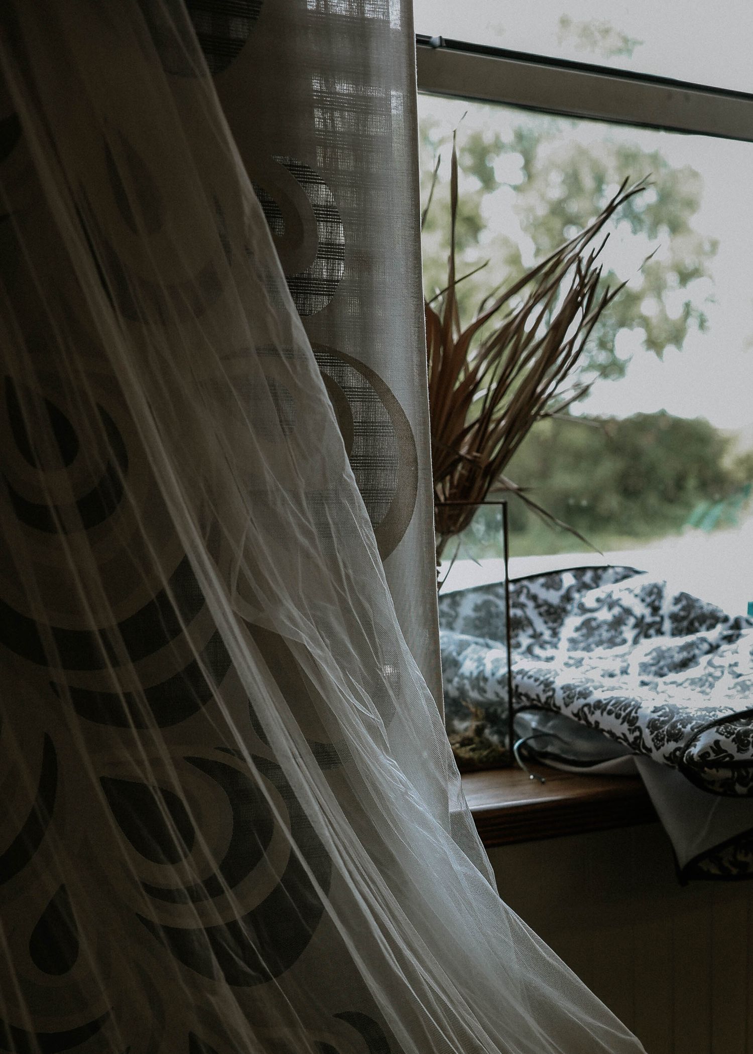 Sheer white curtain billowing near a window with dried plants on a wooden sill and green trees outside.