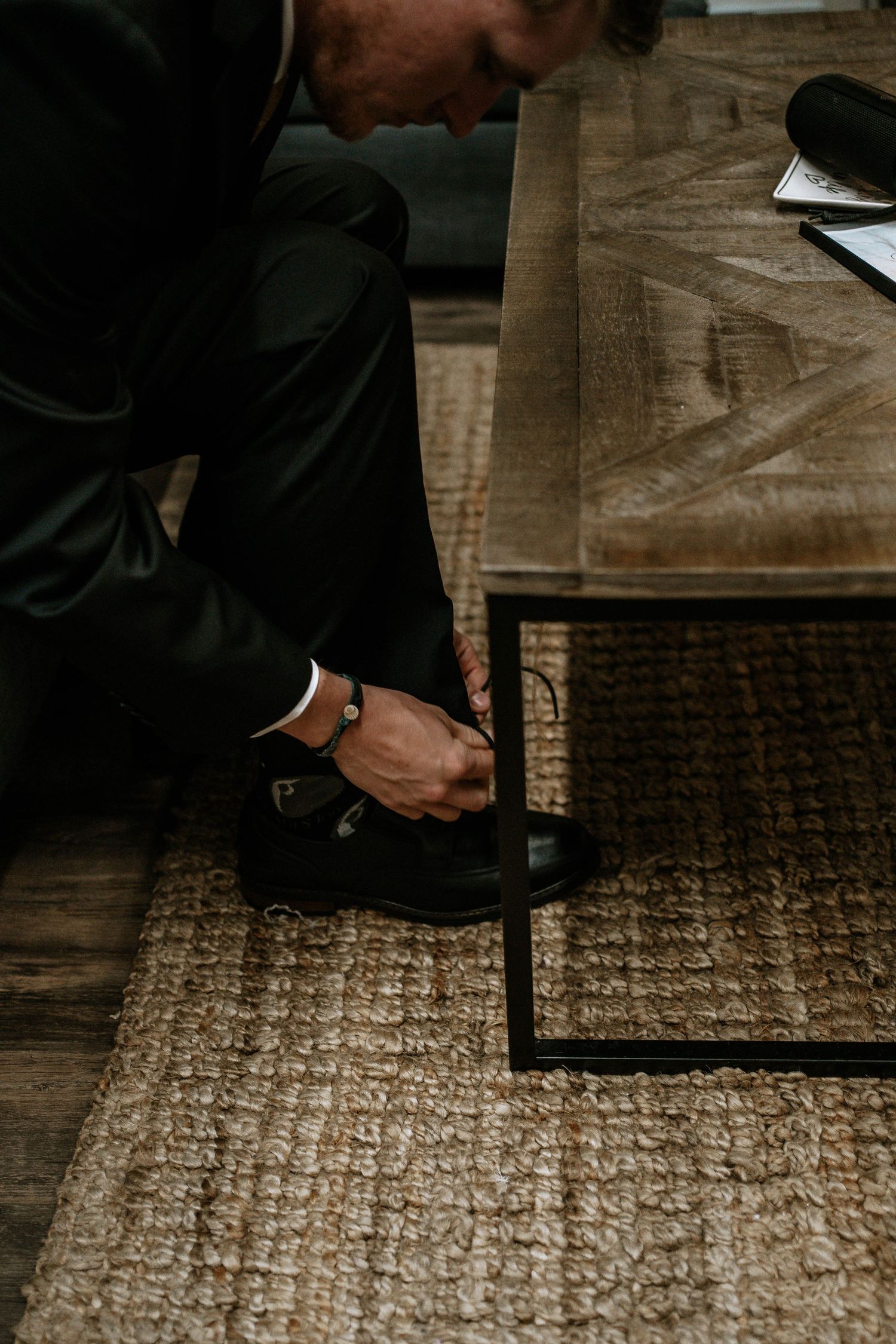 Man in black suit tying his dress shoes on a woven rug beside a rustic wooden coffee table.