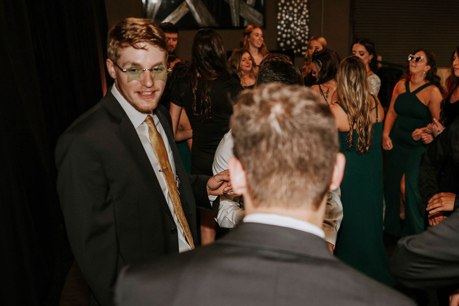 Young man in suit and gold tie smiling at wedding reception with guests in formal attire in background.