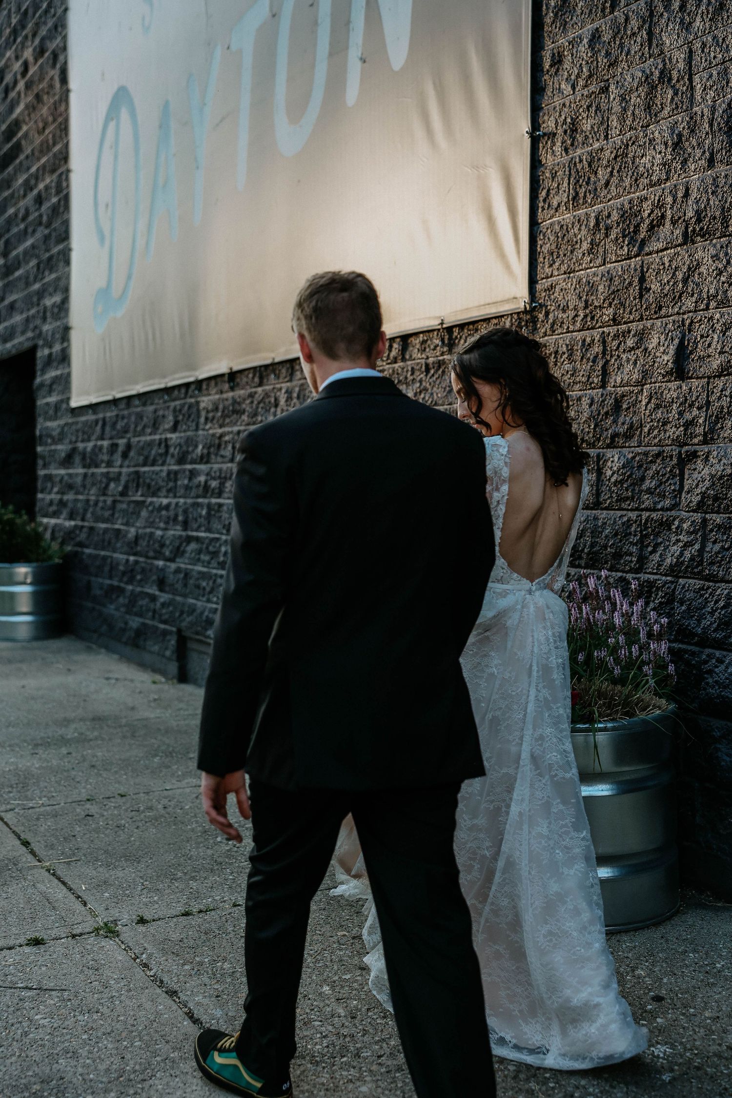 Bride and groom walking together outside a brick building with a Dayton sign, bride wearing a backless lace wedding gown.