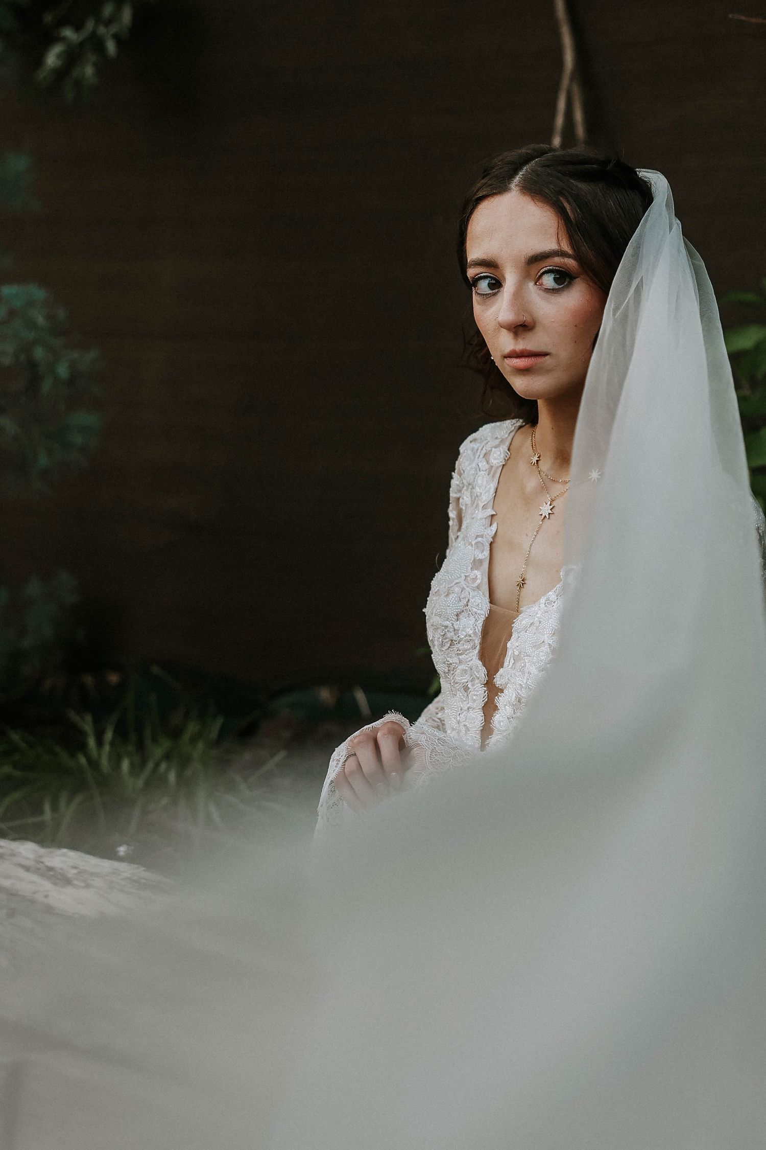 Bride in lace deep-V wedding gown and veil poses outdoors with a contemplative gaze.
