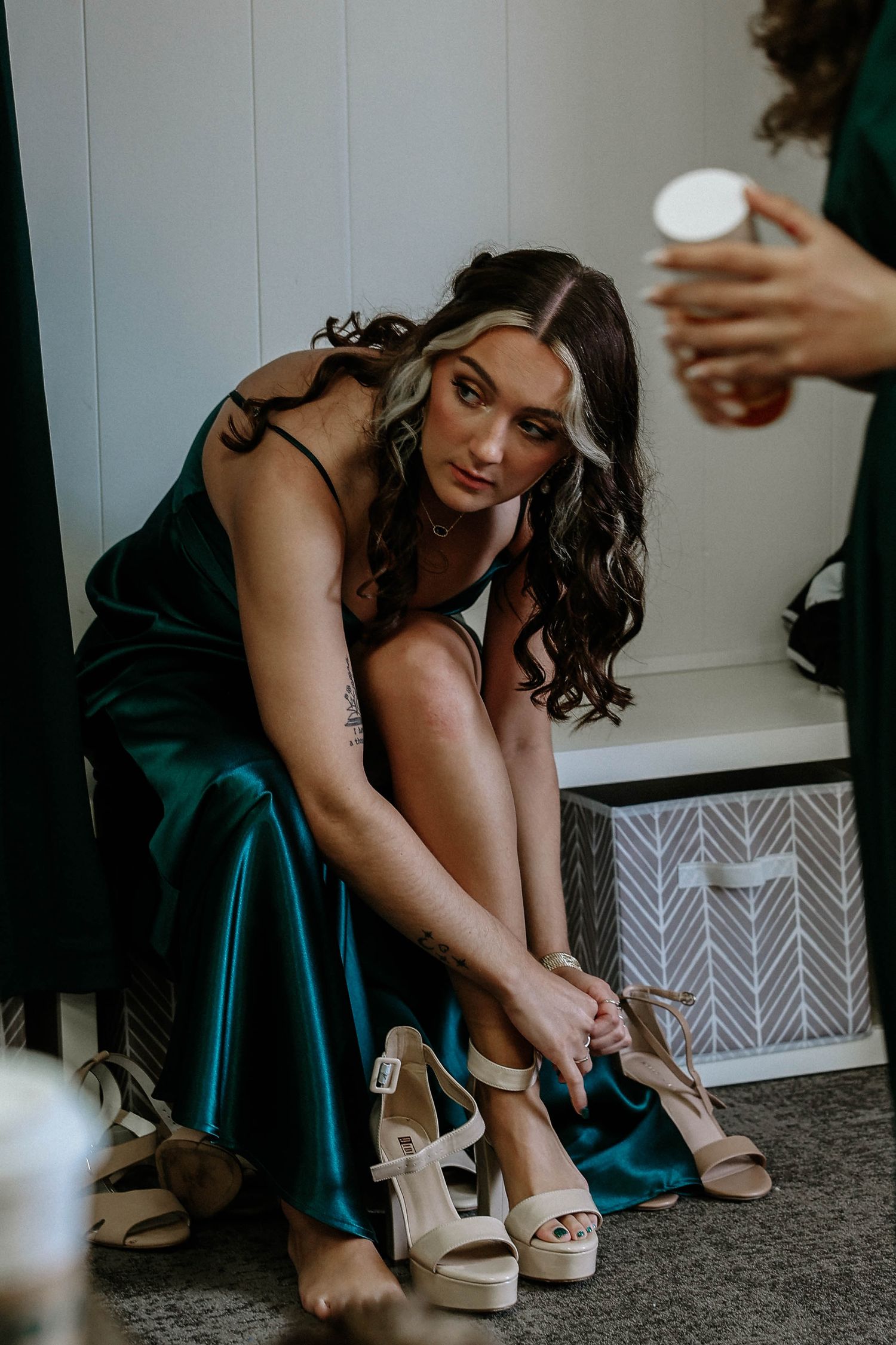 Bridesmaid in emerald green satin dress putting on nude platform heels while sitting on floor.