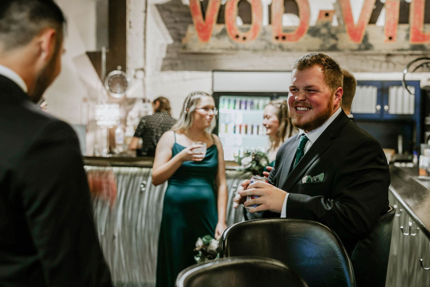 Wedding guests in formal attire laughing and socializing at a bar venue during a wedding reception.