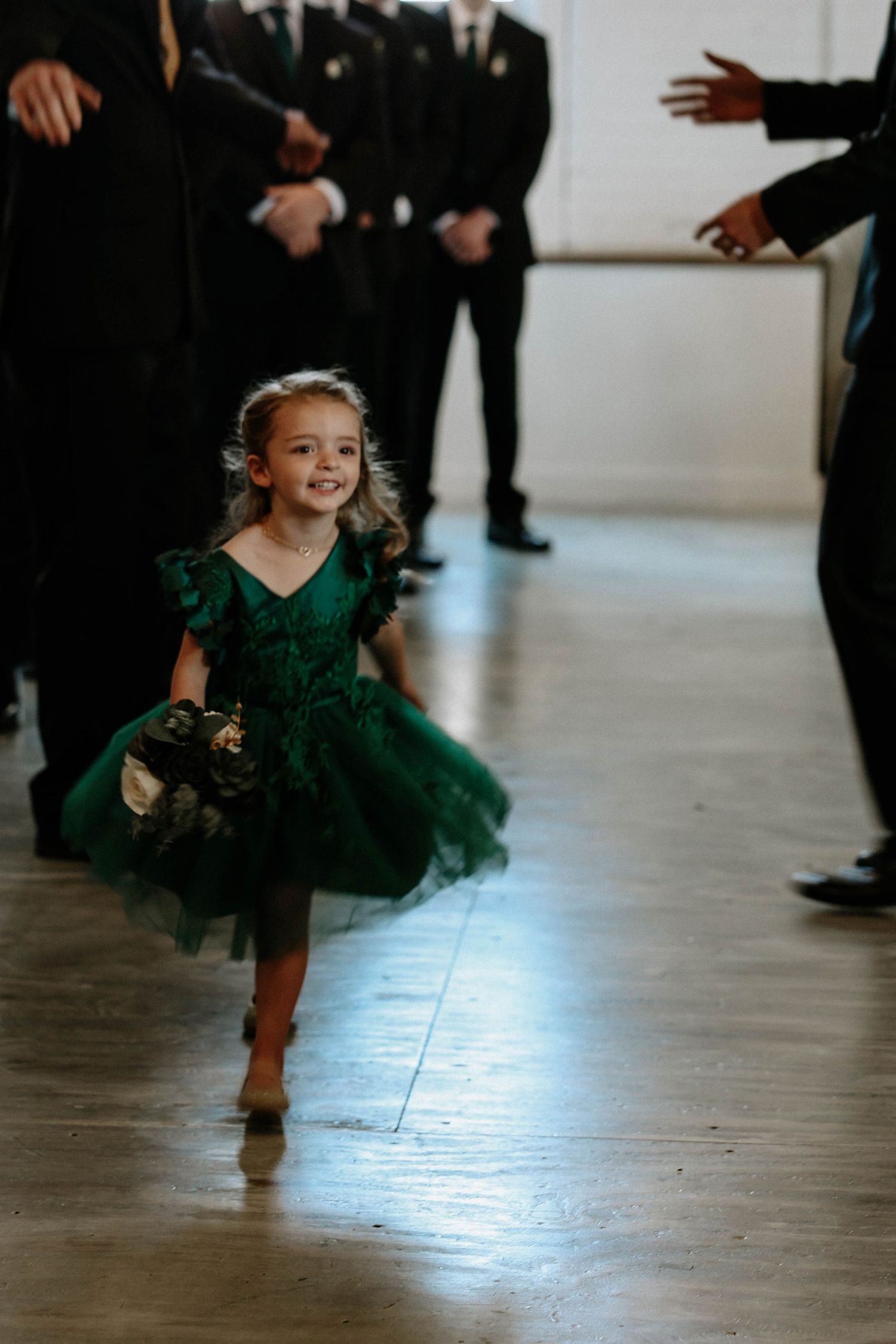 A smiling flower girl in an emerald green tulle dress walks down the aisle at a wedding ceremony.