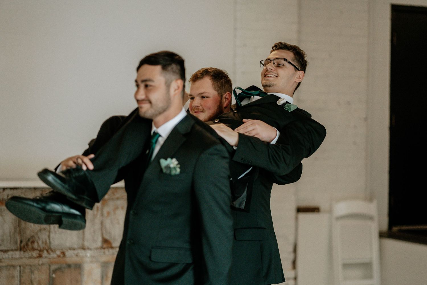 Three groomsmen in dark green suits helping each other get dressed before a wedding ceremony.
