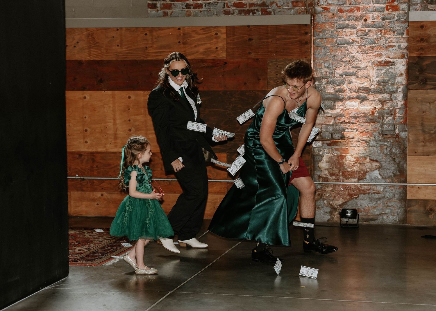 Wedding guests in green and black formal attire tossing money at a lively indoor celebration with brick walls.