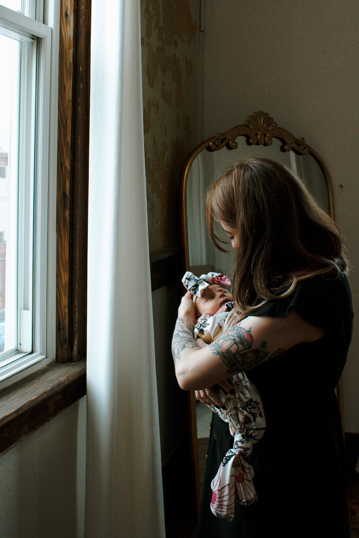 Mother holding newborn baby near window with ornate gold mirror in Cincinnati, Ohio.