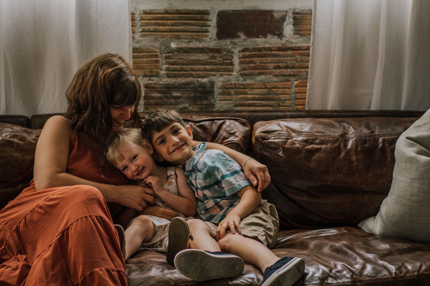 Mother cuddling two young children on a brown leather couch in a cozy living room at Mill Street Studios in Cincinnati, Ohio.