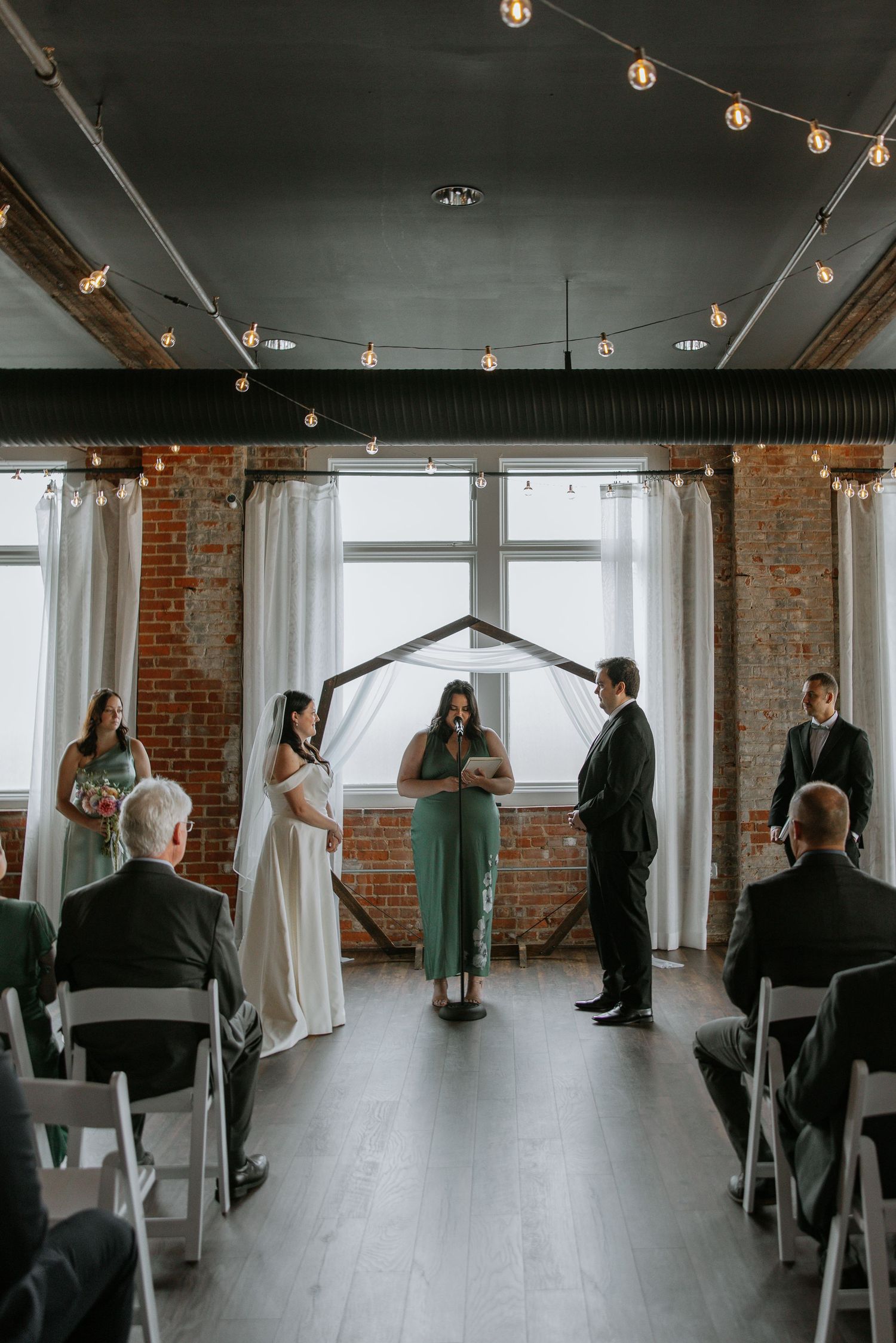 Bride and groom exchange vows at rustic indoor wedding ceremony with string lights and brick walls.