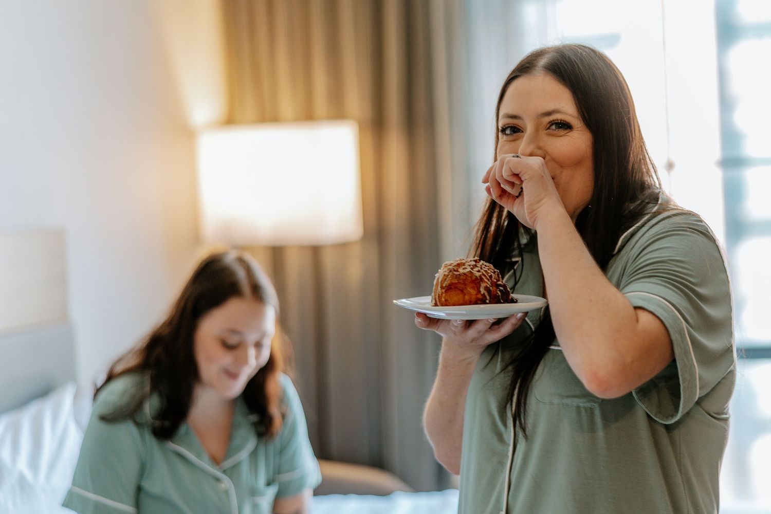 Woman in green pajamas eating a pastry on a plate while another woman sits on the bed behind her.