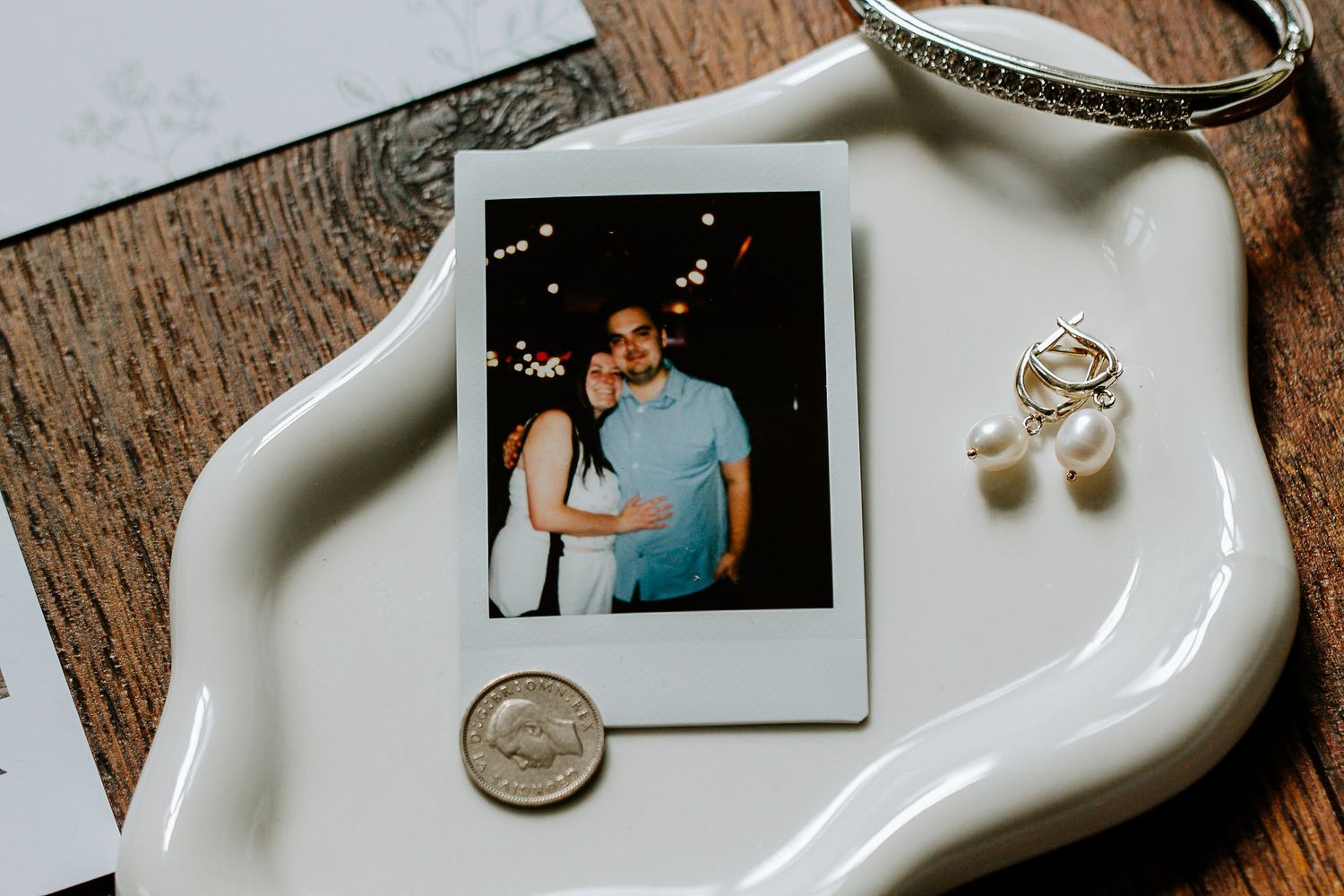 Polaroid photo of couple displayed on white ceramic tray with pearl drop earrings and silver bangle bracelet.