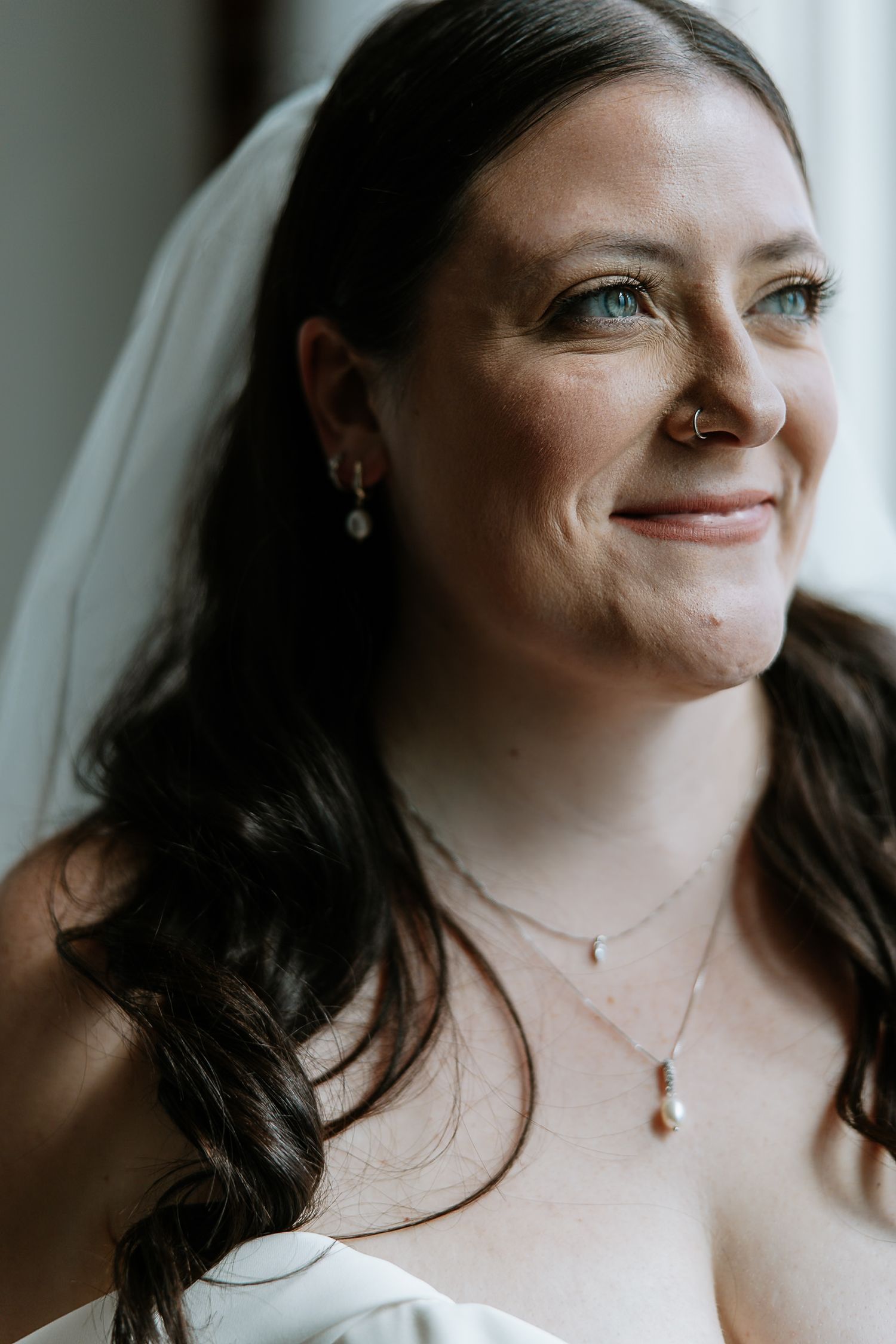 Smiling bride with blue eyes wearing a veil, pearl earrings, and delicate necklace on her wedding day.