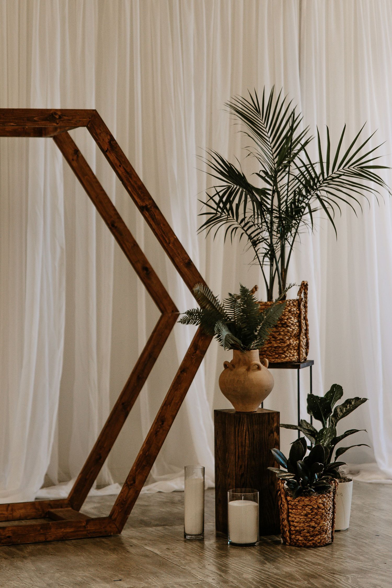 Rustic wooden geometric arch with tropical plants, candles, and wicker baskets against white drape backdrop.