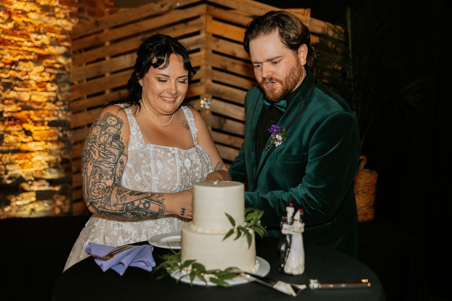 Tattooed bride and groom in green velvet suit cutting white wedding cake together.