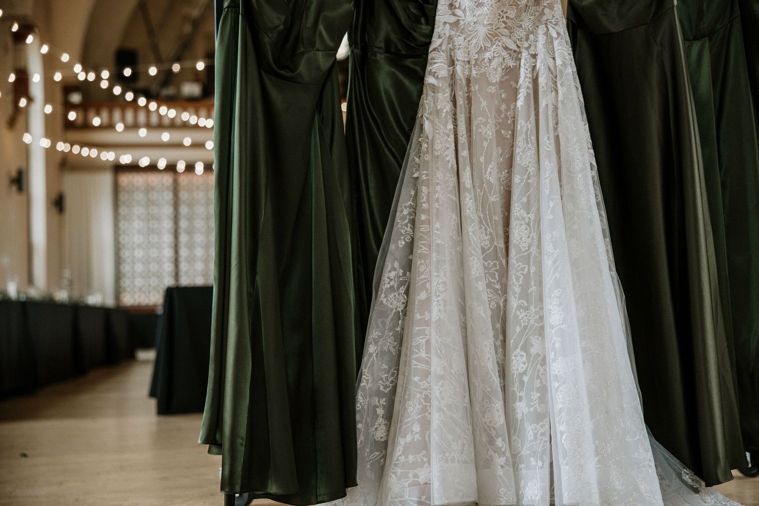Elegant lace wedding gown hanging beside dark green bridesmaid dresses in a softly lit wedding venue at Urban Artifact in Cincinnati, Ohio