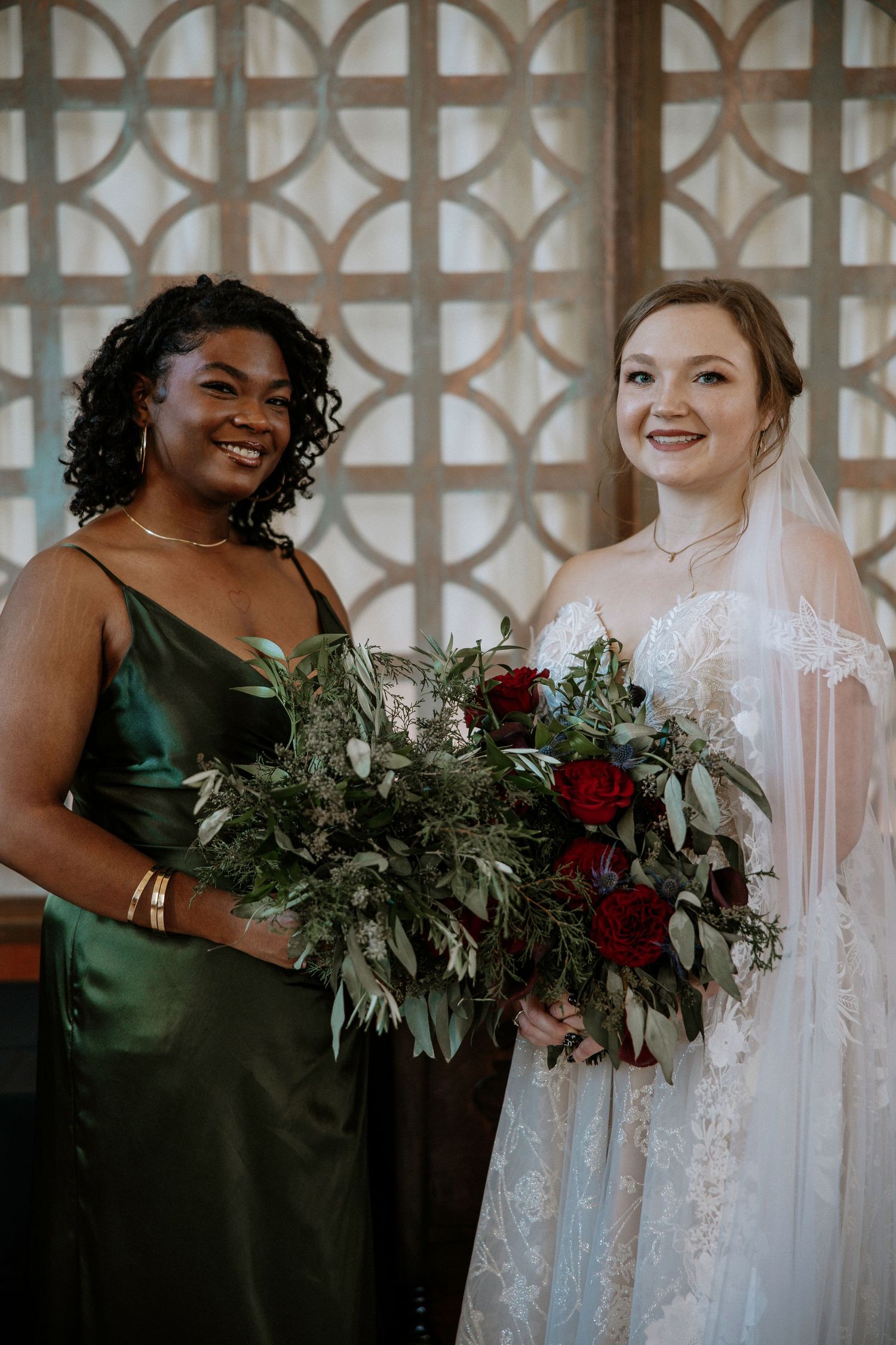 Bride in lace gown holding red rose bouquet poses with bridesmaid in emerald green dress at wedding at Urban Artifact in Cincinnati, Ohio
