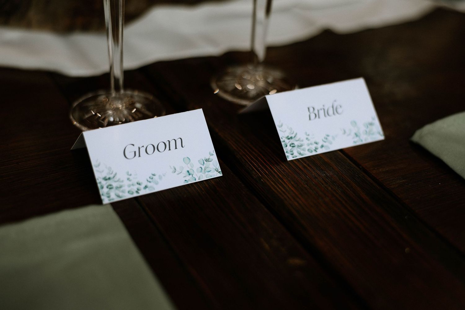 Elegant wedding place cards labeled Groom and Bride with green floral accents on a dark wood table.