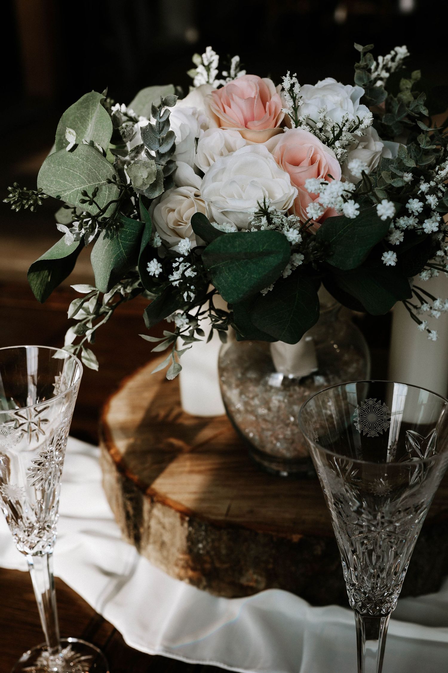 Crystal glasses and a floral centerpiece with pink and white roses on a rustic wood slice at a wedding reception.