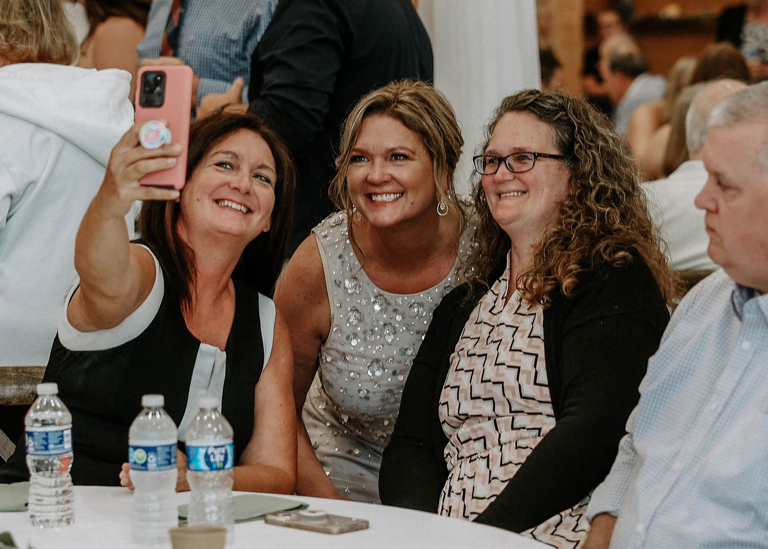 Three women pose for a selfie at a wedding reception, smiling together at a decorated table.