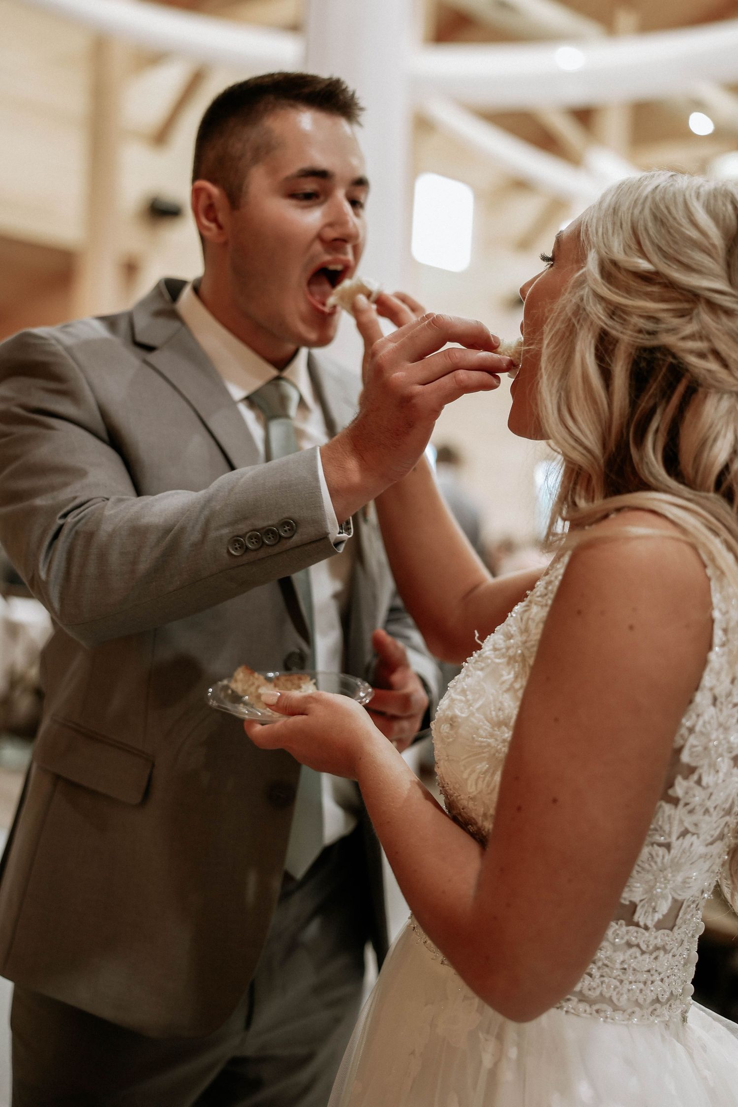 Bride and groom sharing wedding cake during reception, groom in gray suit feeding bride in lace wedding dress.