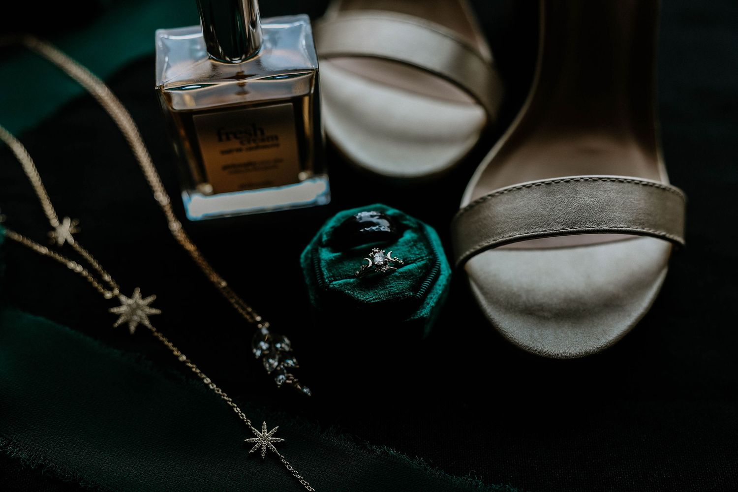 Bridal flat lay with perfume bottle, diamond ring in green velvet box, star necklace, and white strappy heels on dark background.