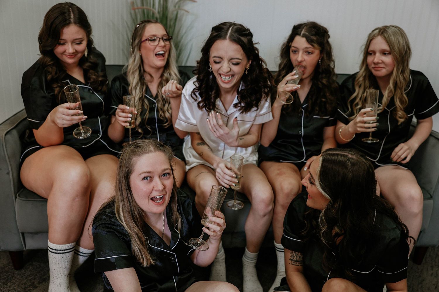 Bridal party in matching black satin pajamas laughing and celebrating with champagne glasses during a fun bachelorette gathering.
