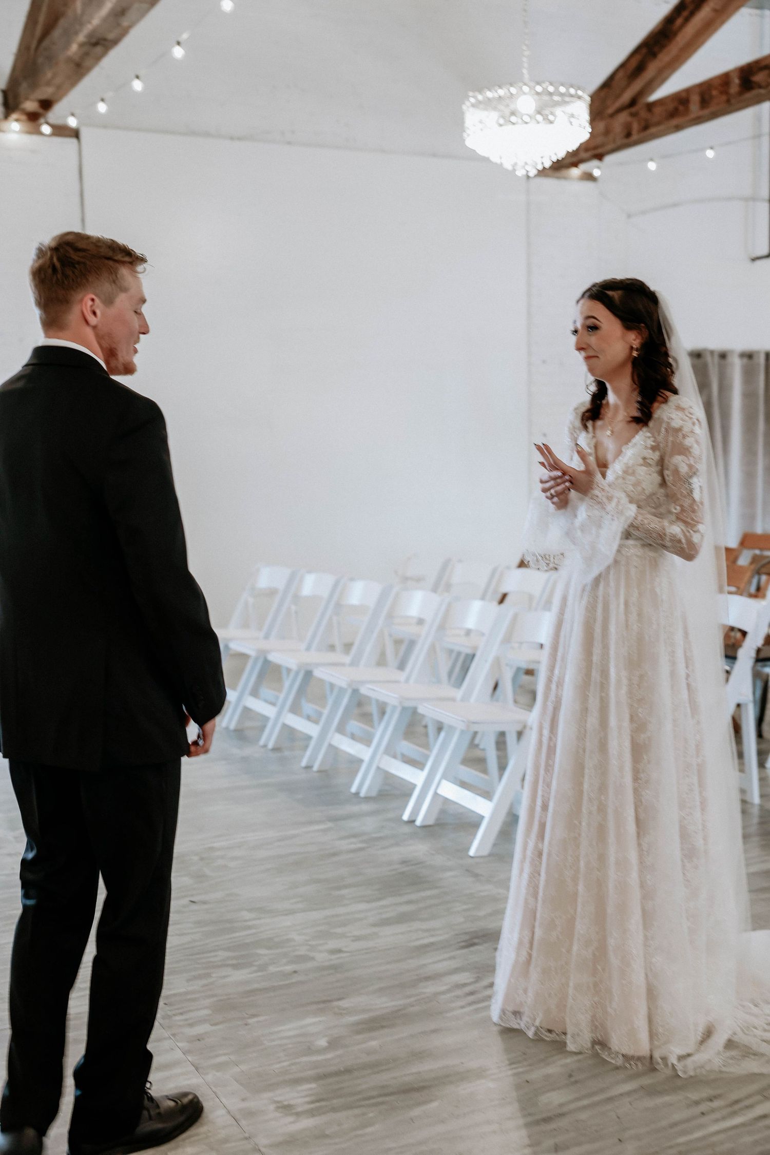 Bride in lace gown and groom in black tuxedo share an intimate first look moment in elegant white wedding venue.