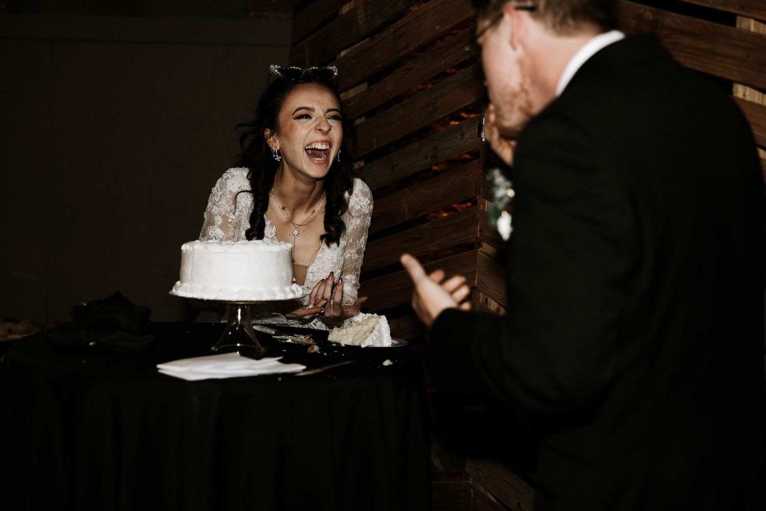 Bride laughing joyfully while cutting white wedding cake at reception, groom in black suit standing nearby.