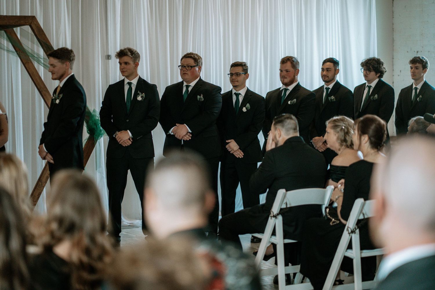 Groomsmen in black suits stand at wedding ceremony altar while guests sit in white chairs.