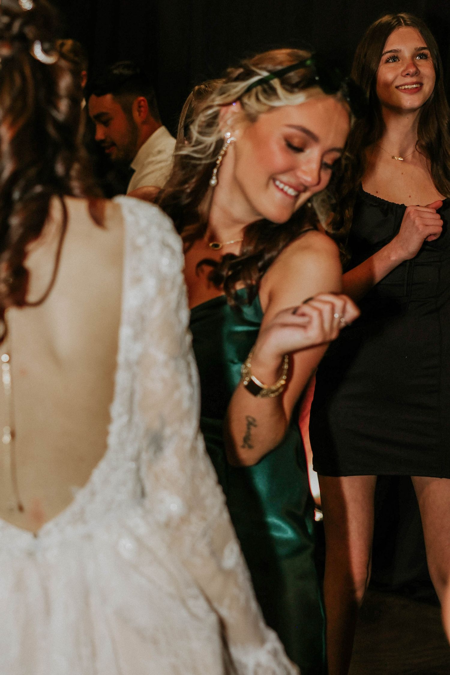 Bride in white lace gown dances joyfully with guests in green and black dresses at a wedding reception.