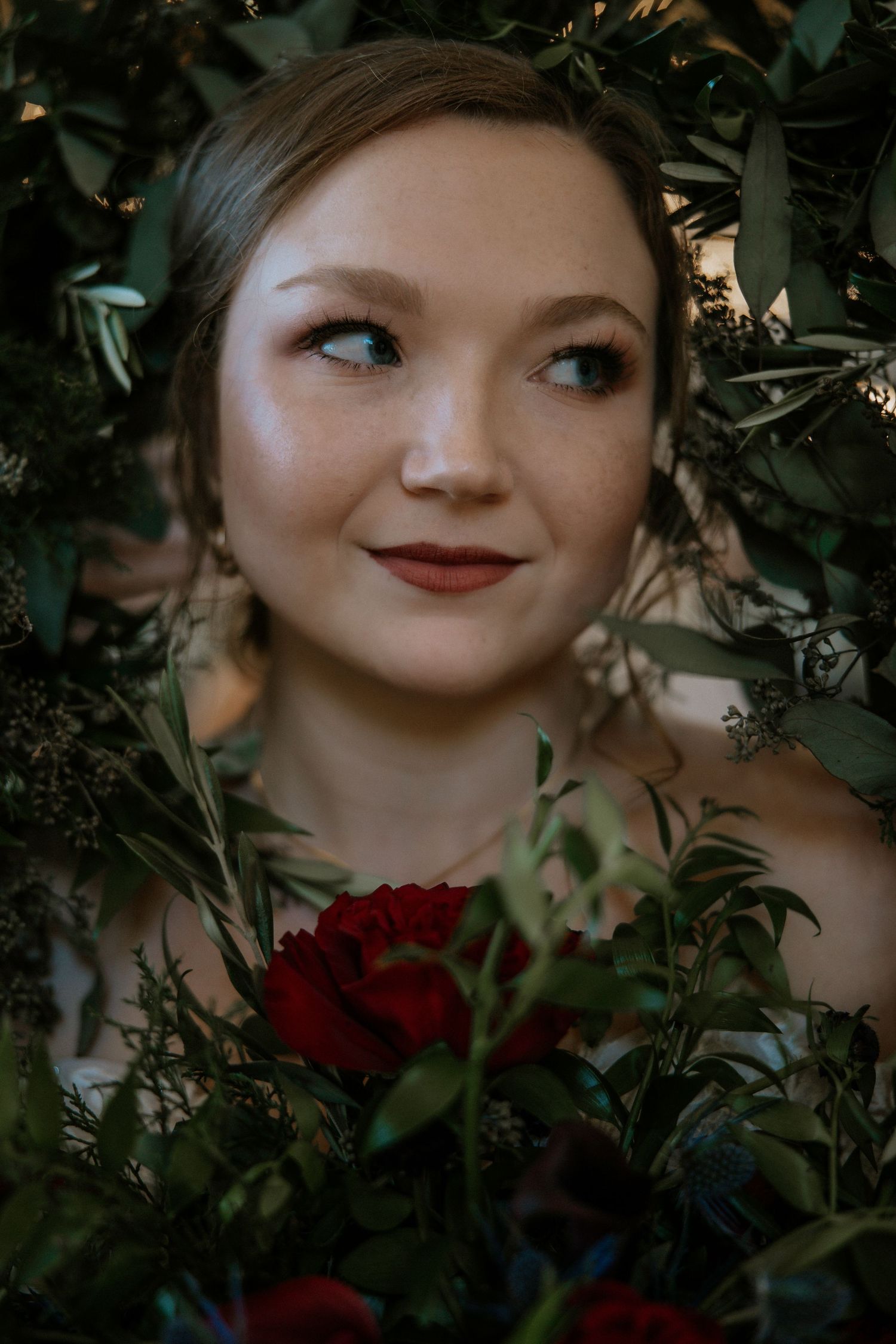 Close-up portrait of a bride with red lips holding red roses surrounded by lush greenery at Urban Artifact in Cincinnati, Ohio