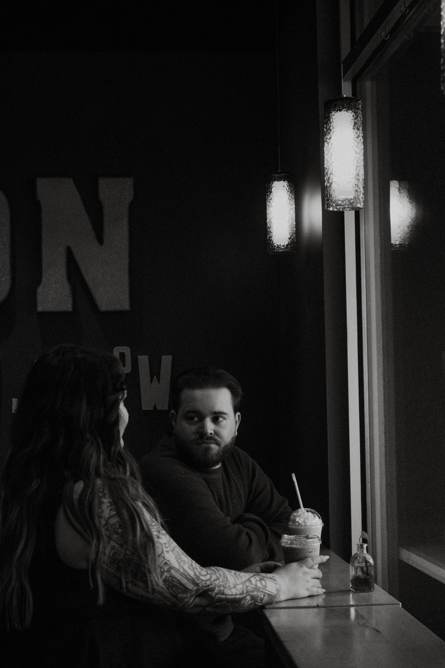 Black and white photo of couple sitting at a dark cafe table by a window with glowing lights in Dayton, Ohio