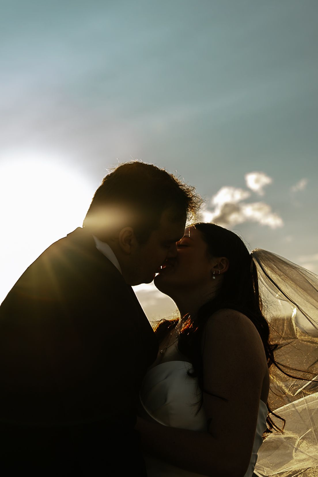 Silhouette of bride and groom kissing at sunset with glowing backlight and veil flowing in wind at The Remedy in Dayton, Ohio