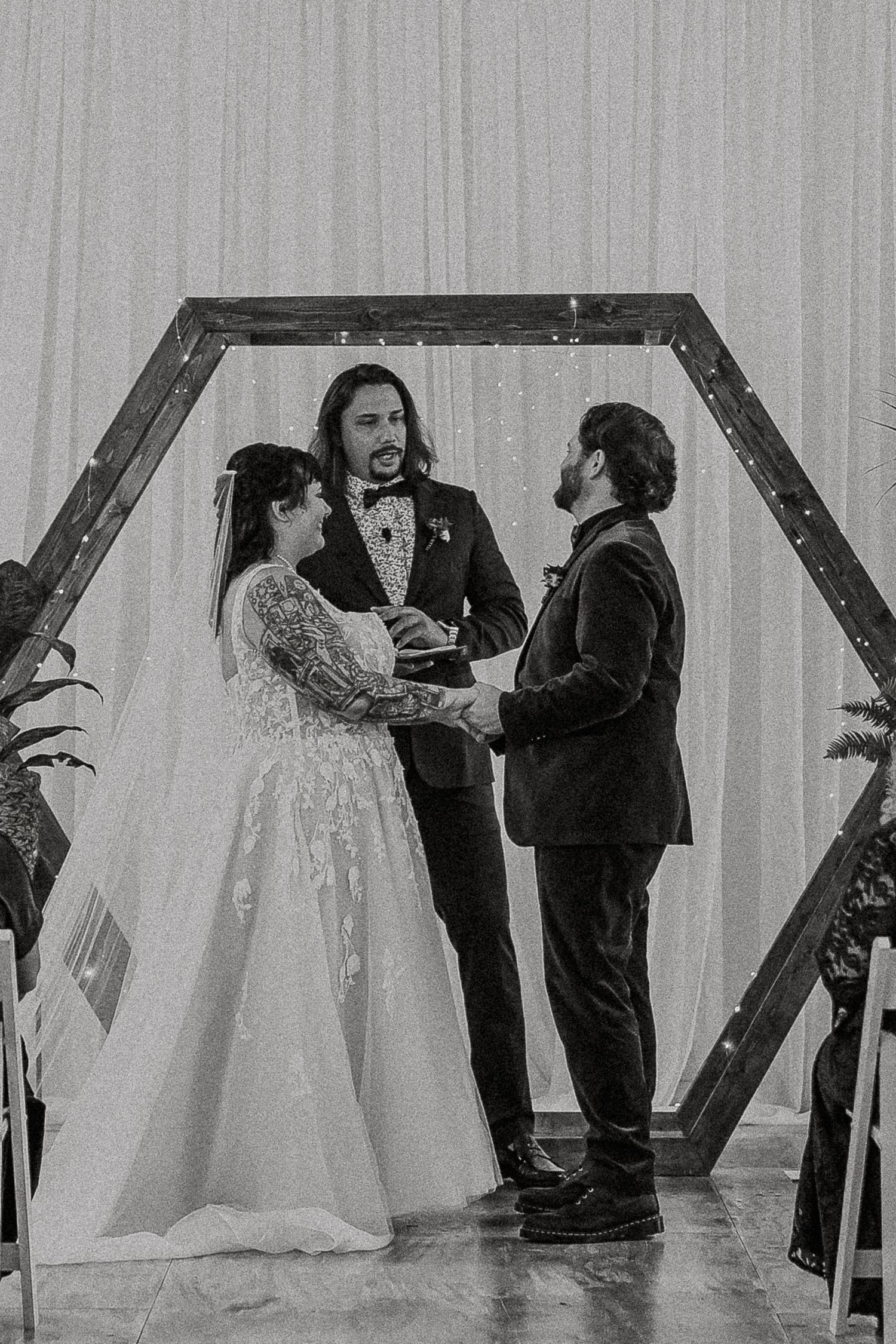 Black and white photo of bride and groom exchanging vows under hexagonal wooden arch.