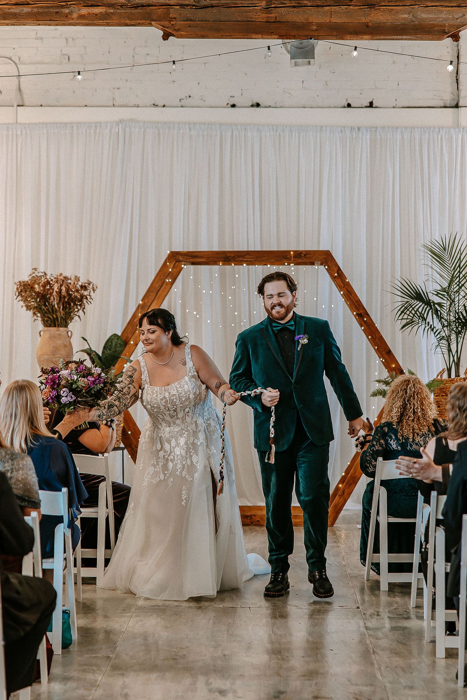 Newlyweds walk joyfully down aisle after ceremony beneath hexagonal wooden arch with twinkling lights.