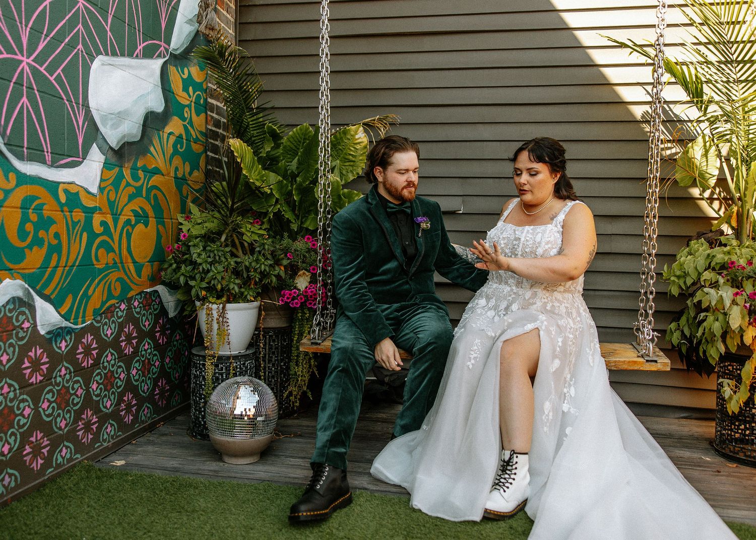 Bride in lace gown with combat boots reads vows to groom in green velvet suit on outdoor swing.