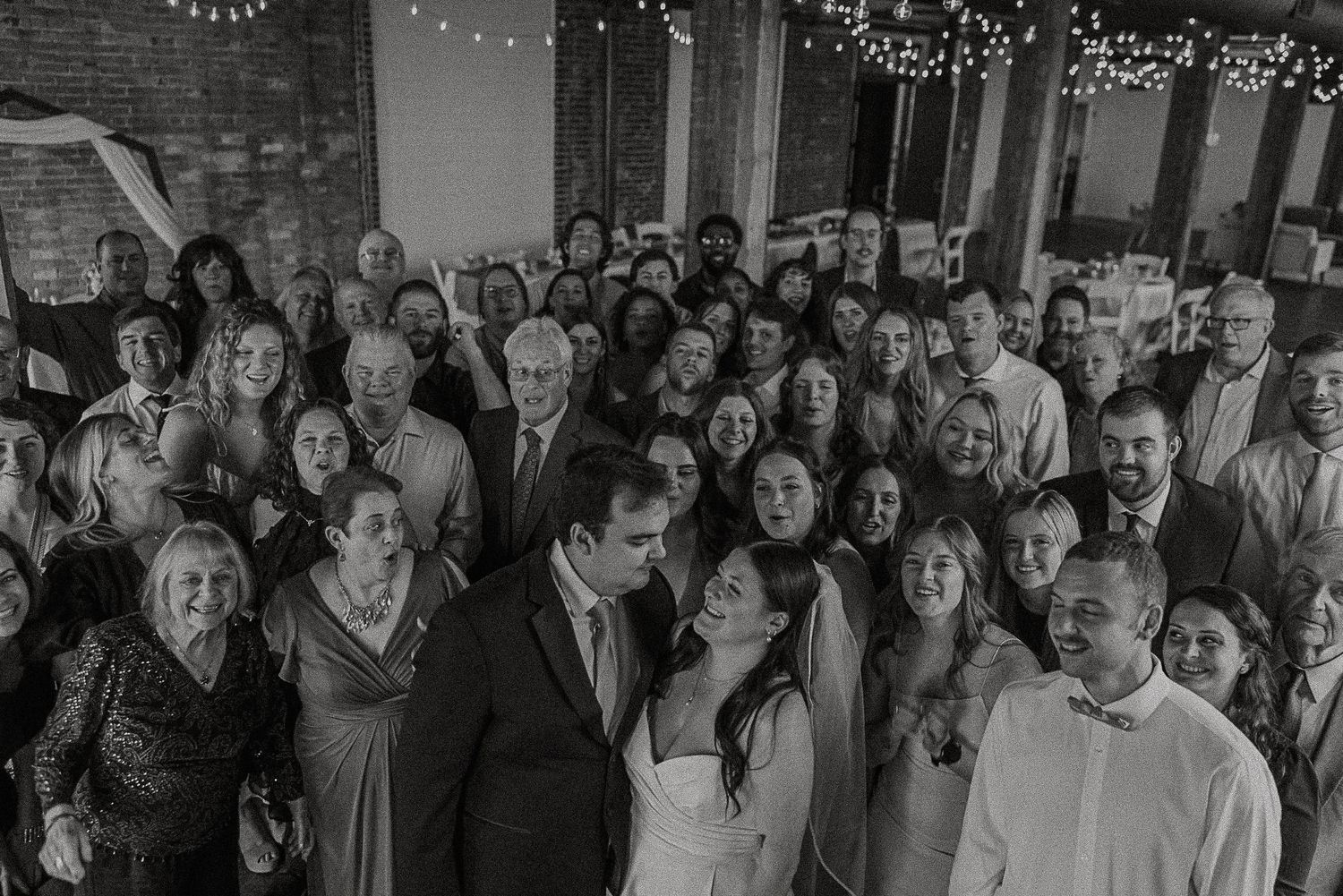 Black and white group photo of wedding couple surrounded by all guests at indoor reception venue.