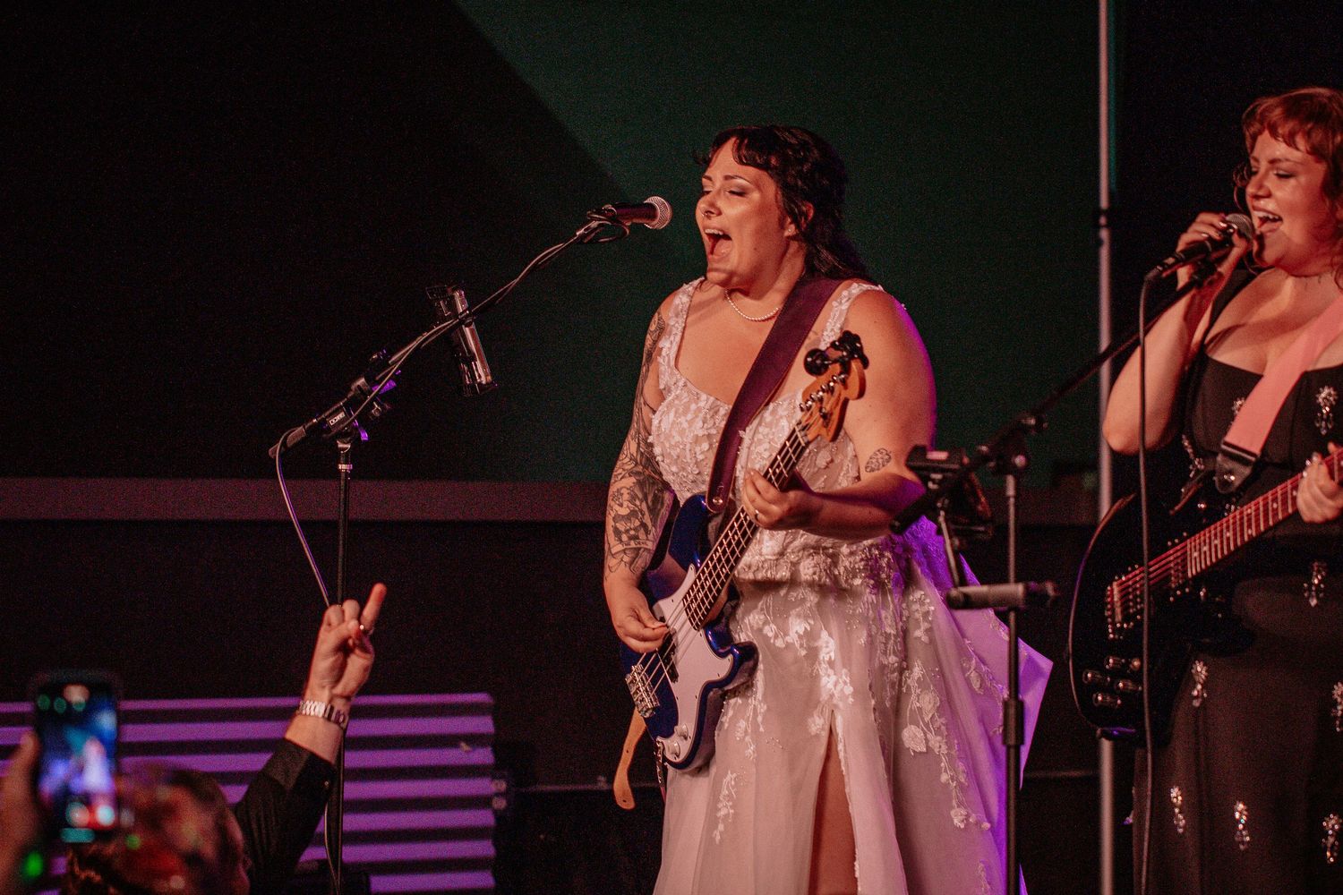 Woman in white dress plays bass guitar and sings on stage at live concert performance.