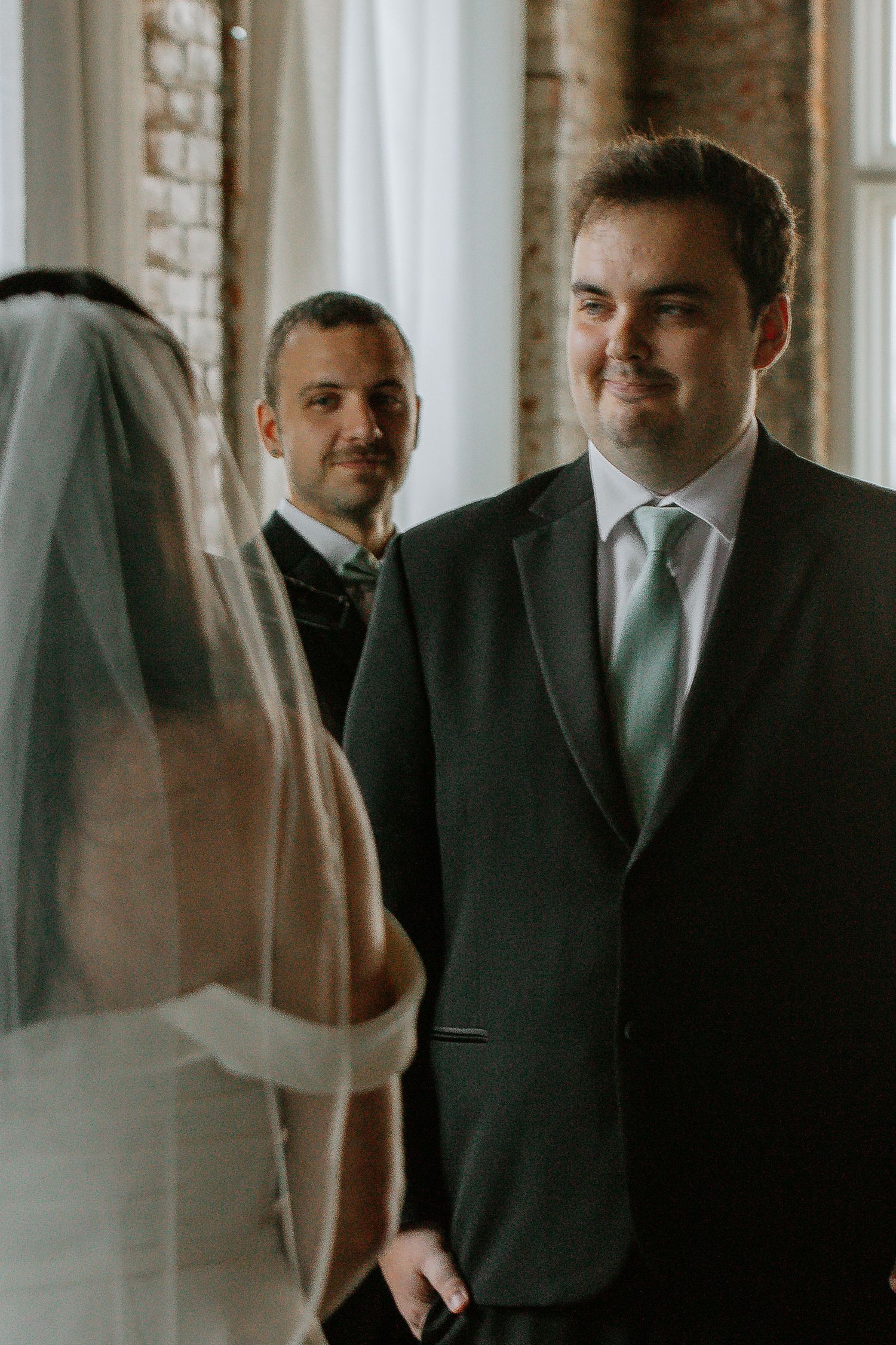 Groom in dark suit with mint tie smiles at bride during wedding ceremony.