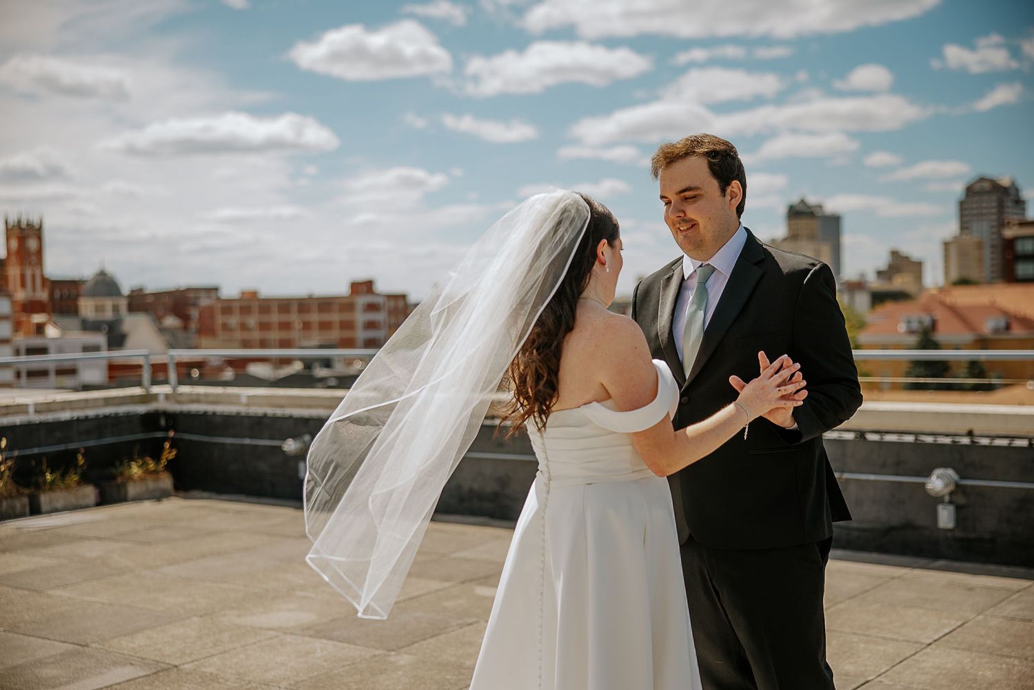 Bride and groom dance on sunny rooftop, bride in off-shoulder gown with veil, city skyline behind.