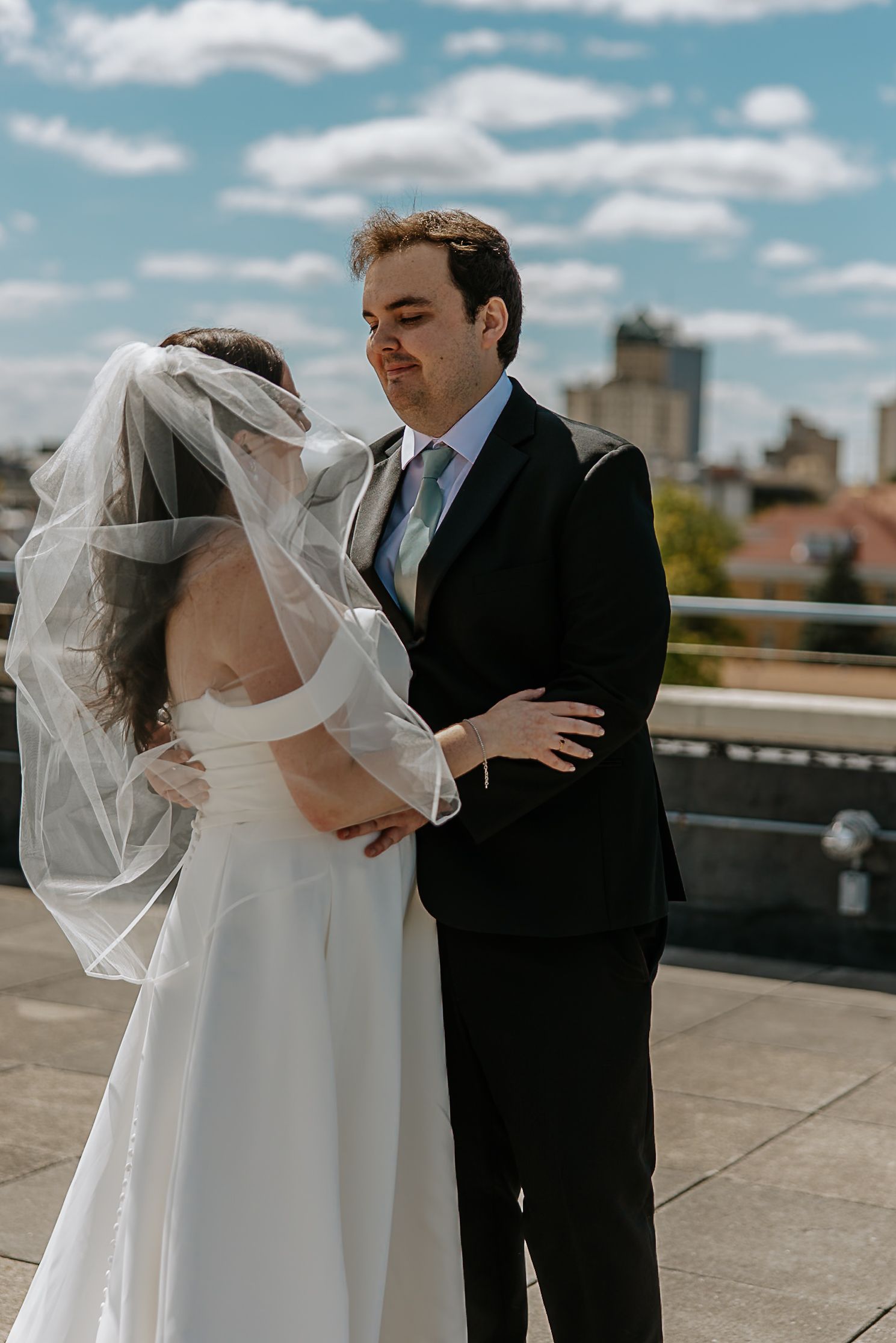 Groom holds bride close on rooftop, her veil blowing in wind, city skyline visible behind them.