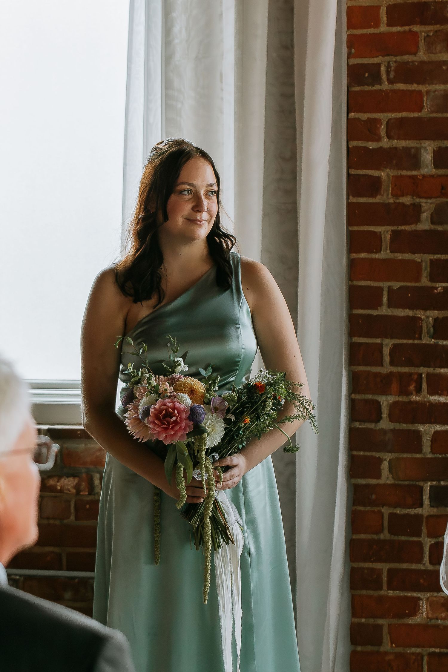 Bridesmaid in sage green gown holding colorful bouquet near brick wall.